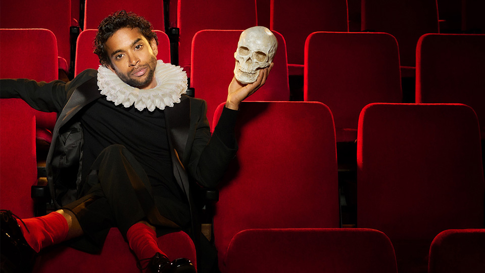 A man wearing a black suit and white collar, sitting on a row of red theatre seats. He is holding a skull in one hand.